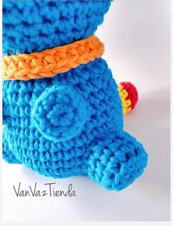 Primer Plano De Un Animal Azul De Crochet Posiblemente Un Oso O Quizás Un Monstruo Colorido Que Muestra La Vista Posterior Con La Cola Y Las Patas Visibles El Animal Presenta Un Collar Naranja Y Un Detalle Rojo Y Amarillo En El Pie Derecho El Texto vanvaztienda Está En La Esquina Inferior Izquierda Lo Que Sugiere Que Podría Ser Un Patrón Gratuito