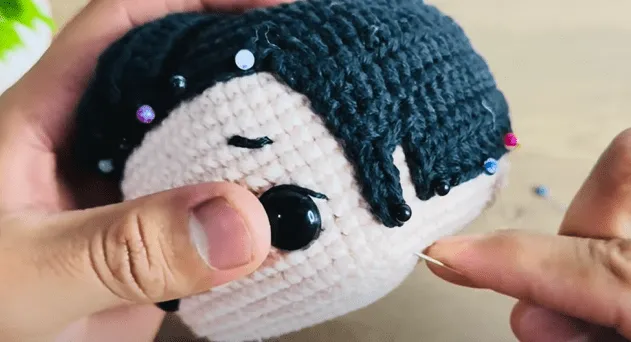Primer plano de manos tejiendo el cabello de una muñeca amigurumi, asegurándolo con alfileres.
