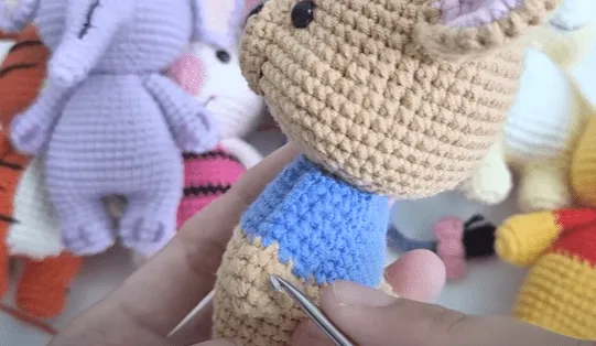 Primer plano de manos tejiendo un amigurumi de Pooh, agregándole una camisa azul.