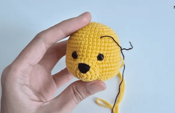 Primer plano de una cabeza de amigurumi amarilla tejida a crochet, con ojos y nariz negros e hilo negro adjunto.