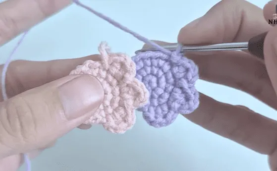 Primer plano de manos tejiendo dos pequeños motivos en forma de flores en hilo rosa y morado.