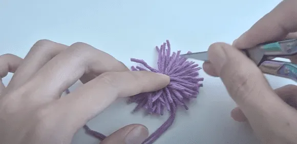 Primer plano de unas manos recortando un pompón de hilo morado.