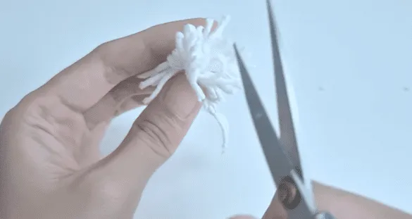 Primer plano de una mano recortando un pompón de hilo blanco con unas tijeras.