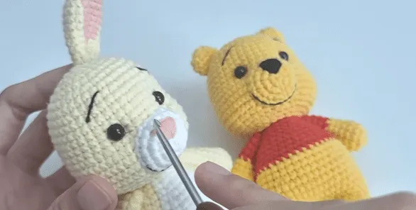 Primer plano de peluches de Winnie the Pooh y conejo tejidos a crochet; la aguja agrega detalles a la nariz del conejo.