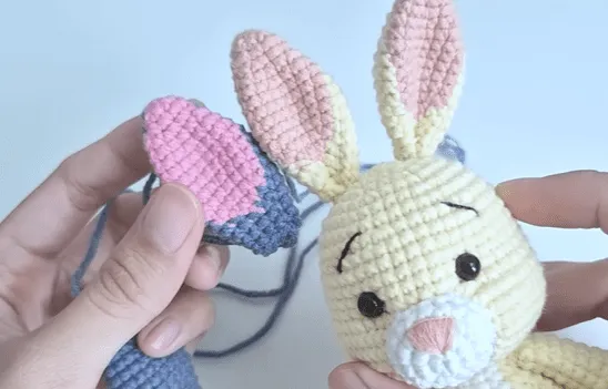 Primer plano de unas manos uniendo orejas de conejo tejidas a crochet a un conejito amigurumi de color pastel.