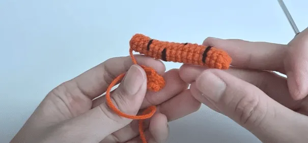 Primer plano de manos tejiendo a crochet una pieza de amigurumi con rayas naranjas y negras.