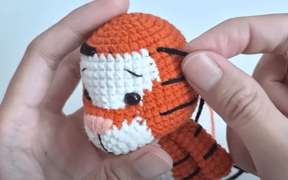 Primer plano de manos tejiendo las rayas de un tigre en un juguete amigurumi.