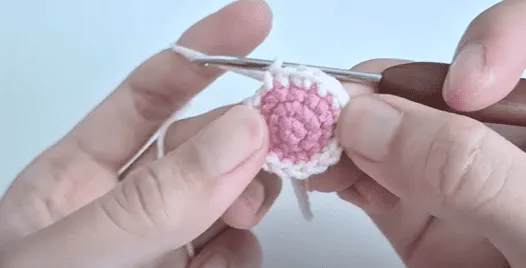 Primer plano de manos tejiendo un círculo amigurumi rosa y blanco.