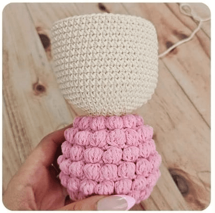 Una Persona Sosteniendo Una Figura De Un Muñeco Parcialmente Tejido a Crochet Usando El Patrón Amigurumi La Parte Superior Está Confeccionada En Hilo Beige Mientras Que La Parte Inferior Es De Hilo Rosa Con Un Punto De Pompón Texturizado El Fondo Presenta Una Superficie De Madera