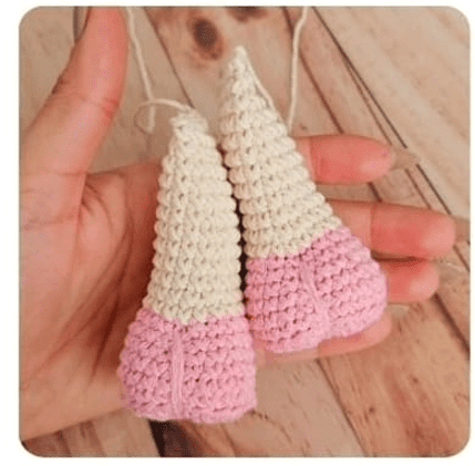 Un Primer Plano De Una Mano Que Sostiene Dos Figuras De Gnomos Tejidas a Crochet Elaboradas Con Técnicas De Crochet Cada Gnomo Es Principalmente Rosa En La Parte Inferior Con Un Sombrero Blanco En Forma De Cono Siguiendo Un Intrincado Patrón Amigurumi El Fondo Es Una Superficie De Madera Clara