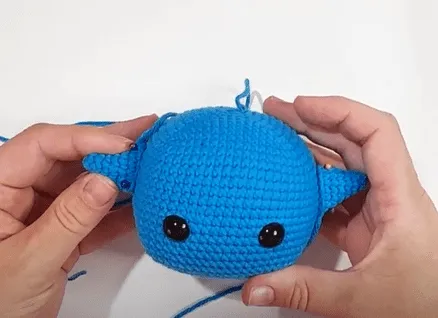Primer plano de manos uniendo orejas de ballena amigurumi tejidas a crochet.