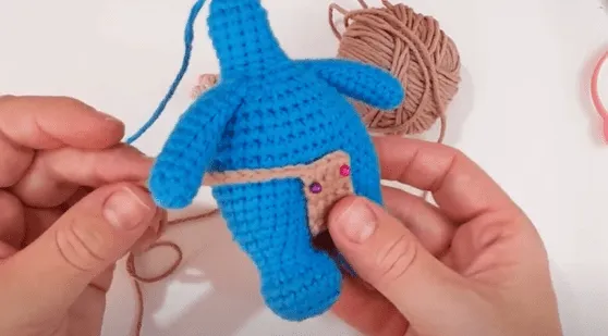 Primer plano de manos tejiendo a crochet un pequeño juguete amigurumi azul con un pañal de tela color canela.