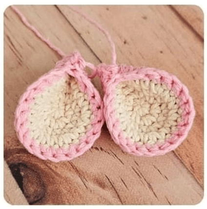 Dos Pequeñas Piezas De Ganchillo Que Se Asemejan a Orejas Se Encuentran Sobre Una Superficie De Madera Cada Pieza Es Principalmente De Color Crema Con Un Borde Rosa Y Tiene Un Hilo Rosa Adjunto Elaborado Usando Técnicas De Crochet De Un Adorable Patrón De Oveja Amigurumi