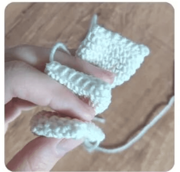 Una Mano Que Sostiene Un Pequeño Proyecto De Crochet Parcialmente Terminado Hecho Con Hilo Blanco Los Puntos Son Claramente Visibles Y El Hilo Está Ligeramente Retorcido Sobre Una Superficie De Madera Hay Un Encantador Alfiletero De Margarita Que Complementa Esta Tranquila Escena