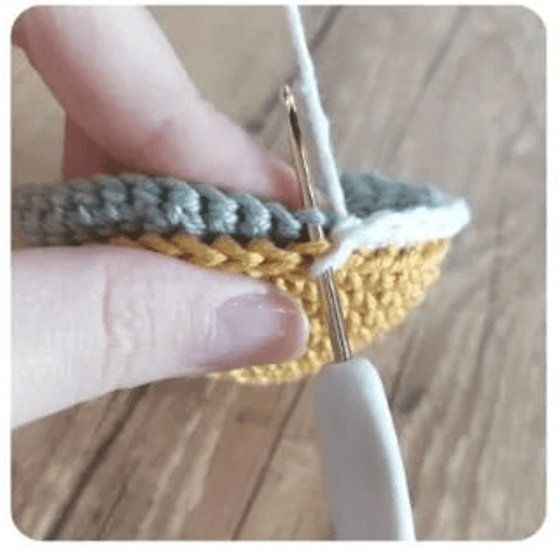 Primer Plano De Una Persona Tejiendo Sosteniendo Un Trozo De Tela Parcialmente Tejido a Crochet El Trabajo De Crochet Utiliza Hilo Gris Y Amarillo Con Un Ganchillo Blanco Insertado En Los Puntos Los Dedos De La Persona Son Visibles Mientras Trabaja En El Proyecto Tal Vez Siguiendo Un Patrón De Costura Gratis El Fondo Muestra Una Superficie De Madera