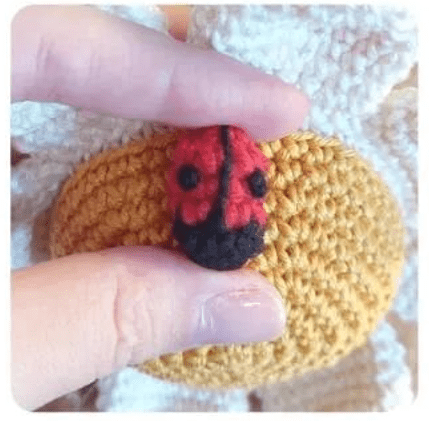 Foto De Primer Plano De Una Mano Sosteniendo Una Pequeña Mariquita De Ganchillo La Mariquita Tiene El Cuerpo Rojo Y Manchas Negras Al Fondo Se Ve Parte De Un Objeto De Crochet Amarillo Más Grande Con Elementos De Crochet Blancos Que Se Asemeja a Un Alegre Alfiletero De Margarita