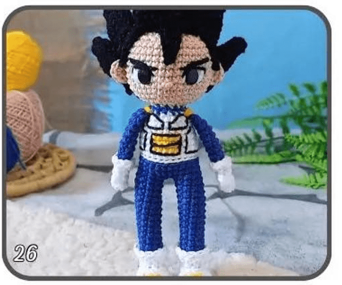 Un Muñeco De Ganchillo Que Se Parece a Un Amigurumi Vegeta Con Cabello Negro Puntiagudo Vestido Con Un Traje Azul Y Blanco Con Detalles En Amarillo Y Guantes Blancos El Fondo Muestra Un Ovillo De Lana Un Ganchillo Y Algo De Vegetación El Número 26 Está En La Esquina Inferior Izquierda De La Imagen