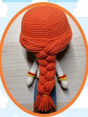 Una Muñeca De Ganchillo Con Pelo Largo Trenzado De Color Naranja Y Guantes Blancos La Muñeca Se Ve Desde Atrás Y Está Enmarcada Por Un Borde Naranja Ovalado El Fondo Presenta Un Cielo Azul Claro Con Nubes Blancas