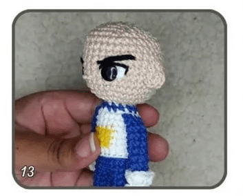 Una Mano Sostiene Un Pequeño Muñeco Amigurumi Con La Cabeza Calva Cejas Prominentes Y Un Traje Azul Con Una Estrella Amarilla En El Pecho El Número 13 Es Visible En La Esquina Inferior Izquierda De La Imagen Mostrando Uno De Los Personajes De Dragon Ball En Crochet