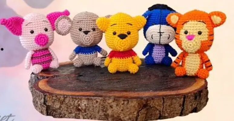 Cinco Pequeños Animales De Peluche Tejidos a Crochet Que Se Asemejan a Personajes De La Serie Winnie the Pooh Están Sentados Sobre Una Plataforma Redonda De Madera De Izquierda a Derecha Un Amigurumi Piglet En Rosa Eeyore En Gris Con Camiseta Azul Pooh En Amarillo Con Camiseta Roja Eeyore En Azul Y Tigger En Naranja Con Rayas perfecto Para Los Entusiastas De Las Manualidades Con Hilo