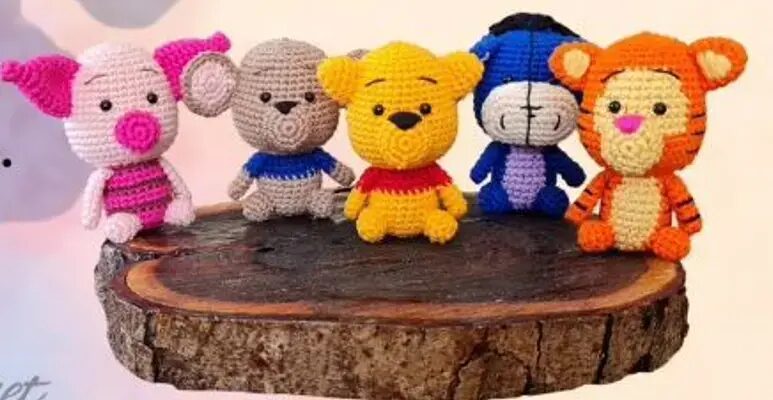 Figuras Tejidas a Mano De Cinco Personajes Familiares Y Coloridos Están Sentadas Sobre Una Losa De Madera De Izquierda a Derecha Un Cerdo Rosa Un Elefante Gris Un Oso Amarillo Que Se Parece a Winnie the Pooh Un Burro Azul Y Blanco Y Un Tigre Naranja Todos Están Creados Con Hilo De Un Patrón De Crochet Y Tienen Una Apariencia Linda Y Caprichosa
