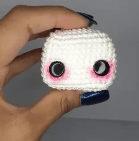 Crea Tu Propio Sombrerero Amigurumi: Patrón Gratuito Paso a Paso en español - Una mano con esmalte de uñas azul oscuro sostiene un pequeño cubo tejido a crochet con grandes ojos negros y rubor rosado en las mejillas sobre un fondo liso y de color claro. Esta adorable figura, creada siguiendo un patrón amigurumi gratis, muestra el encantador arte de las artesanías hechas a mano.