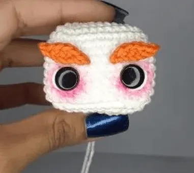 Crea Tu Propio Sombrerero Amigurumi: Patrón Gratuito Paso a Paso en español - Una mano que sostiene un pequeño cubo de crochet hecho a mano con una cara, que recuerda al amigurumi de Alicia en el País de las Maravillas. El cubo presenta grandes ojos blancos y negros, cejas anaranjadas y rubor rosado en las mejillas.