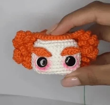 Crea Tu Propio Sombrerero Amigurumi: Patrón Gratuito Paso a Paso en español - Un primer plano de una mano que sostiene un pequeño cuadrado tejido a crochet. El cuadrado, que recuerda al amigurumi de Alicia en el País de las Maravillas, está diseñado para parecerse a un rostro con grandes ojos negros, cejas pobladas de color naranja y cabello rizado de color naranja. El rostro tiene marcas de rubor rosado en las mejillas y una leve sonrisa.