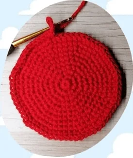 Un Primer Plano De Un Círculo Rojo Tejido a Crochet En Progreso Apoyado Sobre Una Superficie De Madera Se Coloca Un Ganchillo Dorado Con Un Mango Negro En La Parte Superior Del Círculo Y Se Enrolla Un Hilo De Hilo Rojo En El Ganchillo El Fondo Presenta Nubes Blancas Sobre Un Cielo Azul