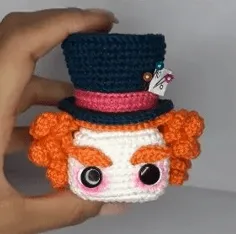 Crea Tu Propio Sombrerero Amigurumi: Patrón Gratuito Paso a Paso en español - Una mano sostiene una figura tejida a crochet del Sombrerero Loco, que presenta un sombrero de copa oscuro con una banda rosa, ojos grandes, cabello rizado naranja y cejas rojas. Directamente sacado del estilo amigurumi de Alicia en el País de las Maravillas, el sombrero del sombrerero está adornado con una pequeña etiqueta y alfileres.