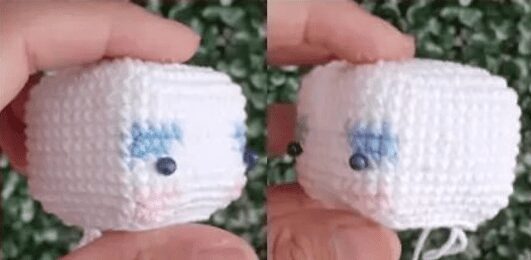 Primer Plano De Un Pequeño Cubo Tejido a Mano Con Hilo Blanco El Cubo Presenta Ojos De Cuentas Negras Cejas Cosidas En Azul Y Un Rubor Rosado En Las Mejillas Se Sostiene Entre Los Dedos Demostrando Su Forma Tridimensional Inspirado En El Amigurumi Alicia En El País De Las Maravillas El Fondo Es Un Follaje Verde
