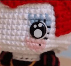 Un Primer Plano De Una Muñeca Amigurumi De Alicia En El País De Las Maravillas Con Un Notable Ojo Grande En Blanco Y Negro Cabello Rojo Y Rubor Rosado En Su Cara Blanca Los Intrincados Puntos De Crochet Y Los Coloridos Detalles Le Dan a La Muñeca Una Apariencia Caprichosa Y Hecha a Mano