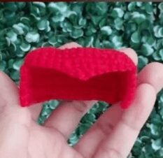 Una Vista De Cerca De Una Persona Sosteniendo Un Pequeño Objeto Rojo Tejido a Crochet Que Se Asemeja a Un Pequeño Sofá El Fondo Se Compone De Follaje Verde Y Frondoso El Intrincado Diseño Alude a Los Patrones Amigurumi De Alicia En El País De Las Maravillas