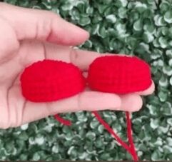 Primer Plano De Una Mano Que Sostiene Dos Pequeñas Piezas De Ganchillo Redondas Y Rojas Frente a Un Follaje Verde Las Piezas Están Conectadas Por Una Hebra De Hilo Rojo Que Se Asemeja Al Crochet Reina De Corazones De Los Cuentos Amigurumi De Alicia En El País De Las Maravillas