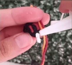 Primer Plano De Manos Pegando Tres Hilos De Colores negro Rojo Y Amarillo Con Una Pistola De Pegamento Caliente De Color Blanco El Fondo Está Borroso Con Follaje Verde Visible perfecto Para Agregar Un Toque De Vitalidad a Tu Proyecto De Crochet Reina De Corazones O Cualquier Patrón Gratuito De Patrón Amigurumi En El Que Estés Trabajando