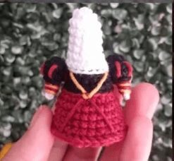 Se Representa Una Pequeña Muñeca Tejida a Crochet Parecida a La Reina De Corazones De Alicia En El País De Las Maravillas Con Un Tocado Alto Blanco Y Un Vestido Rojo Y Negro Con Adornos Dorados El Fondo Está Formado Por Follaje Verde Y La Muñeca Se Sostiene Con Delicadeza En Una Mano