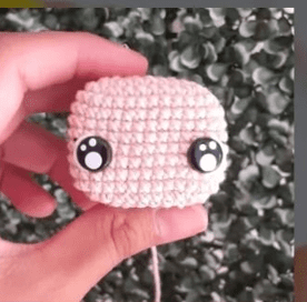 Una Mano Sostiene Una Pequeña Pieza De Crochet Que Se Asemeja a Una Cabeza Con Dos Ojos Redondos En Blanco Y Negro Mostrando Un Adorable Amigurumi De Alicia En El País De Las Maravillas El Fondo Está Borroso Y Parece Ser Una Superficie Texturizada Y Frondosa