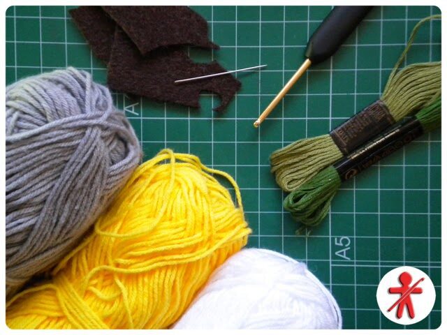 Un Espacio De Trabajo Para Manualidades Presenta Ovillos De Hilo En Gris Amarillo Y Blanco Un Ganchillo Negro Hilo De Bordar Verde Una Aguja De Coser Y Pequeños Trozos De Fieltro Marrón Sobre Una Estera De Corte Verde Con Un Patrón De Cuadrícula En La Esquina Inferior Derecha Hay Un Logotipo Con Una Figura De Palo Rojo