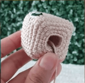 Una Foto De Primer Plano Muestra Una Mano Sosteniendo Un Pequeño Cubo Tejido a Crochet De Color Beige El Cubo Tiene Un Agujero Circular En El Centro De Una Cara El Fondo Está Borroso Con Follaje Verde Es Perfecto Para Quienes Siguen Un Patrón De Amigurumi Gratuito Para Crear Su Propia Colección De Amigurumi Alicia En El País De Las Maravillas