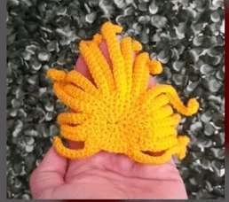 Una Mano Sostiene Una Pieza De Ganchillo Amarilla Con Forma De Pulpo El Fondo Se Compone De Follaje Verde Y Blanco La Pieza Tejida a Crochet Inspirada En Un Patrón Amigurumi De Alicia En El País De Las Maravillas Tiene Tentáculos Extendidos