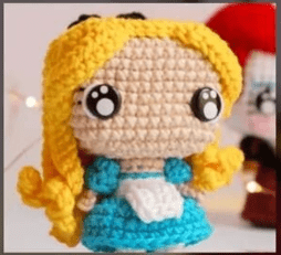 Una Muñeca De Crochet Con Largo Cabello Amarillo Grandes Ojos Redondos Vestida Con Un Vestido Azul Y Un Delantal Blanco La Muñeca Tiene Una Linda Apariencia De Estilo Chibi El Fondo Borroso Incluye Otra Figura Tejida a Crochet Con Cabello Rojo mira El Tutorial De Amigurumi En Español Para Ver Creaciones Adorables Similares