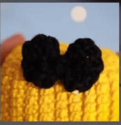 Una Imagen En Primer Plano De Un Objeto Tejido De Color Amarillo Con Un Lazo De Ganchillo Negro Adherido Que Recuerda a Un Amigurumi De Alicia En El País De Las Maravillas El Fondo Es Azul Claro Con Un Pequeño Círculo Blanco Cerca De La Parte Superior Del Marco