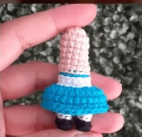 Una Mano Sostiene Una Pequeña Muñeca De Crochet Sin Cabeza Ni Brazos La Muñeca Lleva Una Falda Azul Una Blusa Blanca Y Zapatos Negros El Fondo Es Borroso Lo Que Lo Hace Perfecto Para Exhibirlo En Un Tutorial De Amigurumi En Español