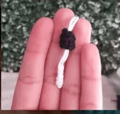 Un Primer Plano De Una Mano Sosteniendo Un Pequeño Hilo Blanco Con Un Nudo Negro Que Recuerda a Los Detalles Encontrados En Un Amigurumi De Alicia En El País De Las Maravillas El Fondo Presenta Follaje Verde Ligeramente Borroso