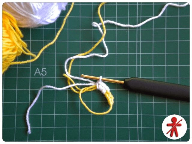 Proyecto De Crochet En Curso Sobre Una Alfombra De Corte Verde Se Utiliza Un Ganchillo E Hilo Blanco Y Amarillo Para Formar Una Cadena Los Carretes De Hilo Son Parcialmente Visibles En La Esquina Superior Izquierda Un Símbolo De Una Persona Tejiendo En Rojo Se Encuentra En La Esquina Inferior Derecha