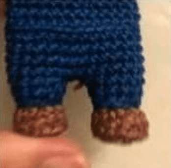 Una Vista De Primer Plano De La Parte Inferior Del Cuerpo Y Las Piernas De Una Muñeca De Crochet Hecha a Mano El Muñeco Elaborado a Partir De Un Patrón Amigurumi Gratuito Viste Unos Pantalones Azules Con Los Pies Hechos De Hilo Marrón Los Dedos De Una Persona Son Visibles En La Parte Inferior De La Imagen Sosteniendo La Muñeca