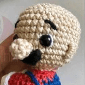 Un Primer Plano De Un Muñeco Amigurumi De Mario Bros Que Se Asemeja a Un Personaje De Dibujos Animados Con La Cabeza Calva Nariz Grande Ojos Negros Y Redondos Espesas Cejas Negras Y Bigote Negro La Muñeca Lleva Un Traje Rojo Y Azul