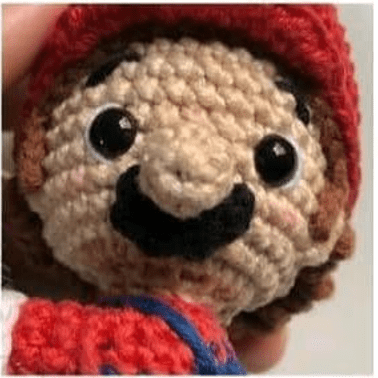 Una Vista De Cerca De La Cara De Un Muñeco Amigurumi De Mario Bros Con Un Tupido Bigote Negro Y Ojos Negros Redondos La Muñeca Lleva Un Sombrero Rojo Y La Artesanía Muestra Costuras Detalladas Lo Que Le Da Al Rostro Un Aspecto Expresivo Y Alegre