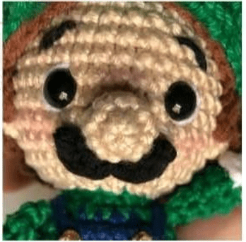 Muñeco De Ganchillo Que Representa Un Personaje Con Un Sombrero Verde La Muñeca Tiene Dos Grandes Ojos Blancos Y Negros Una Nariz Grande Un Bigote Negro Y Viste Ropa Verde Y Azul El Trabajo De Crochet Es Detallado Mostrando Varios Puntos Y Texturas Capturando a La Perfección La Esencia Del Amigurumi Mario Bros