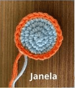 Un Círculo Tejido a Crochet Con Un Centro Azul Y Un Borde Naranja Se Coloca Sobre Un Fondo De Madera La Palabra janela Está Escrita Debajo Del Círculo Dos Hilos De Hilo Cuelgan Del Círculo Asemejándose a Elementos De Un Intrincado Proyecto De Amigurumi Cohete